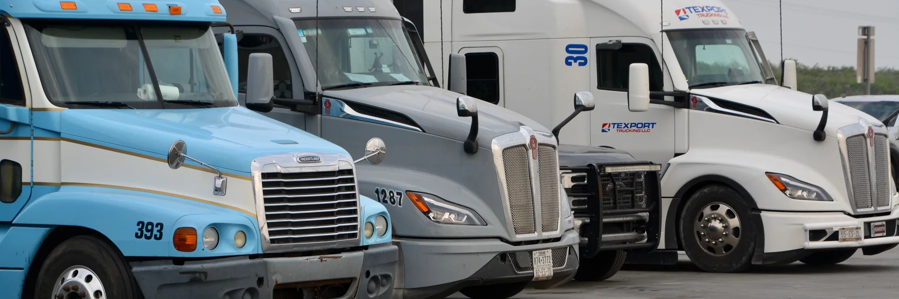 Three conventional semi trucks parked in a flat industrial freight yard.